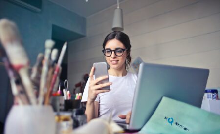 Artist sitting at table looking at cellphone while working on a laptop