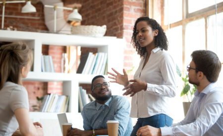Woman standing around a table as her colleagues watch her giving a speech