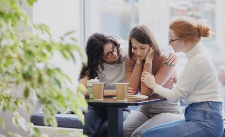 Three women sitting around a table giving support to the woman in the center who is sad
