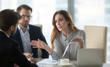 Woman in striped shirt talking to two men seated at a work table